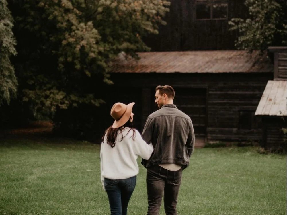 Couple walking on grass near a wooden building with trees in the background, woman wearing a hat and sweater, man in a denim jacket.