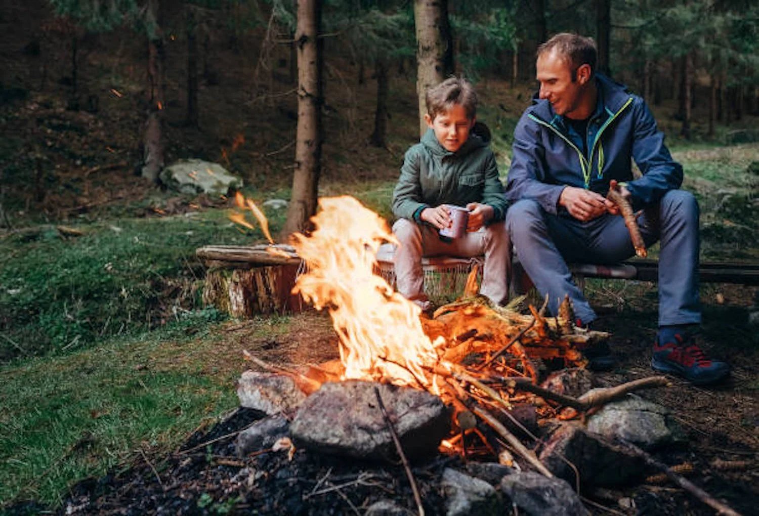 father and young son sitting by a campfire in woods