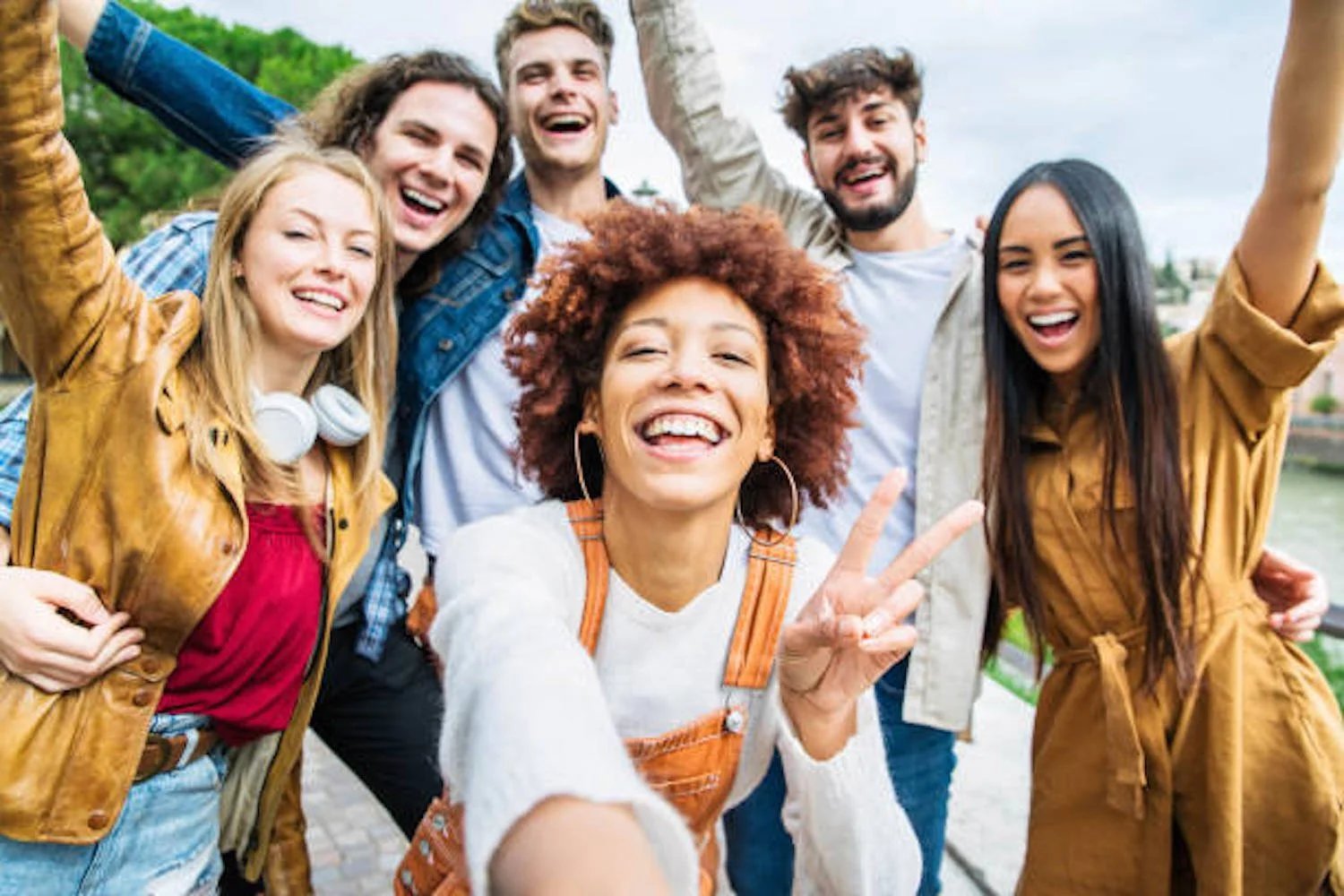 Group of six young adults outdoors, smiling, posing for a selfie, showing excitement and joy, casual attire, trees in the background.